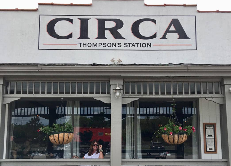 A woman wearing sunglasses sits inside a restaurant with hanging flower baskets and a sign reading 'Circa Thompson's Station' in Williamson County, Tennessee.
