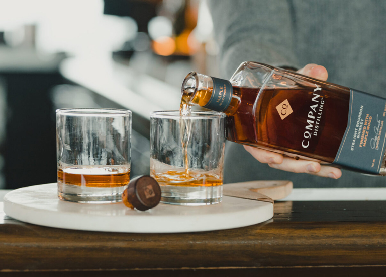 A person pours bourbon from a bottle into two glasses on a wooden bar in Williamson County, Tennessee.