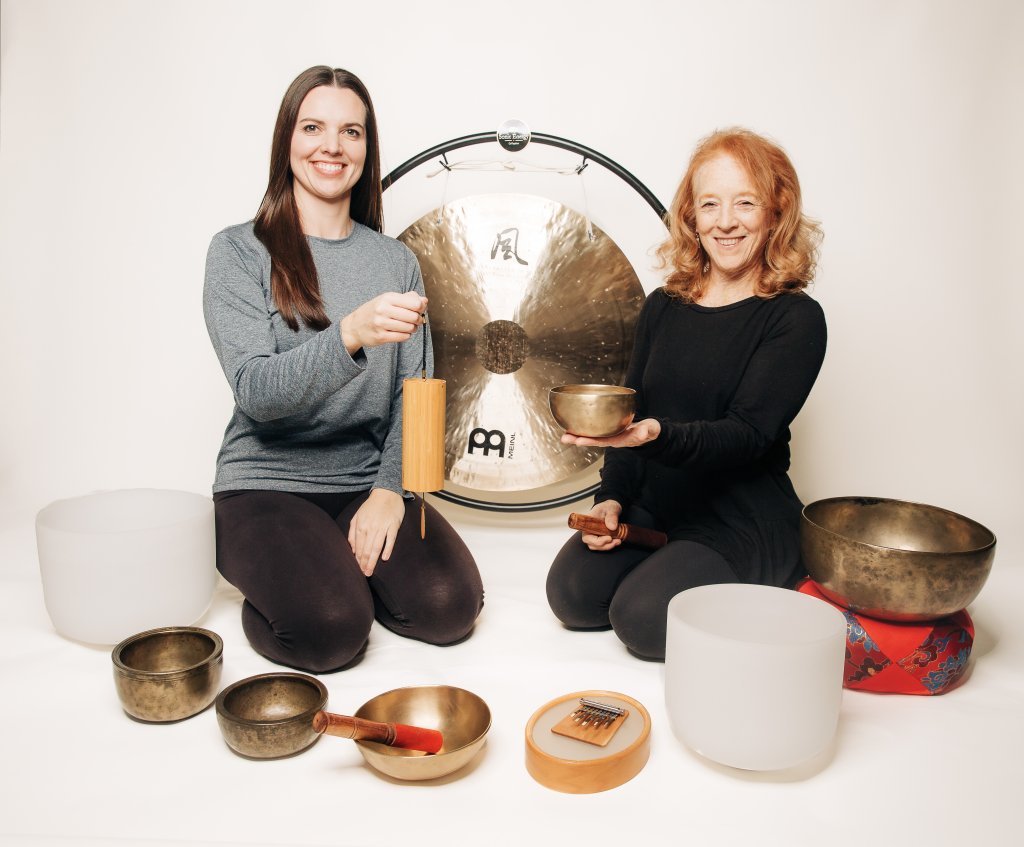 Grounded Vibrations Two women sit cross-legged with singing bowls and a large gong, smiling in Williamson County, Tennessee.