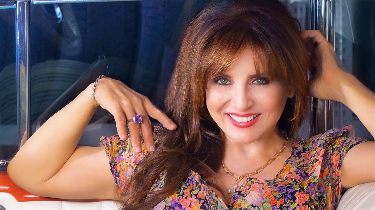 Deborah Allen A woman with long brown hair smiles brightly while wearing a colorful floral dress and jewelry in Williamson County, Tennessee.