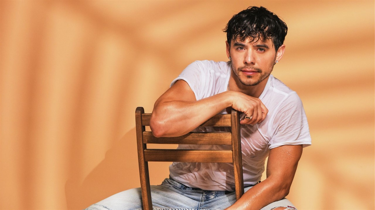 A young man with dark hair and a beard sits casually on a wooden chair, wearing a white t-shirt and jeans in Williamson County, Tennessee.