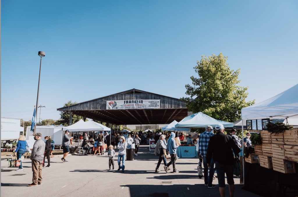 A bustling outdoor market with vendors under tents, people shopping, and a large wooden structure in the background in Williamson County, Tennessee.