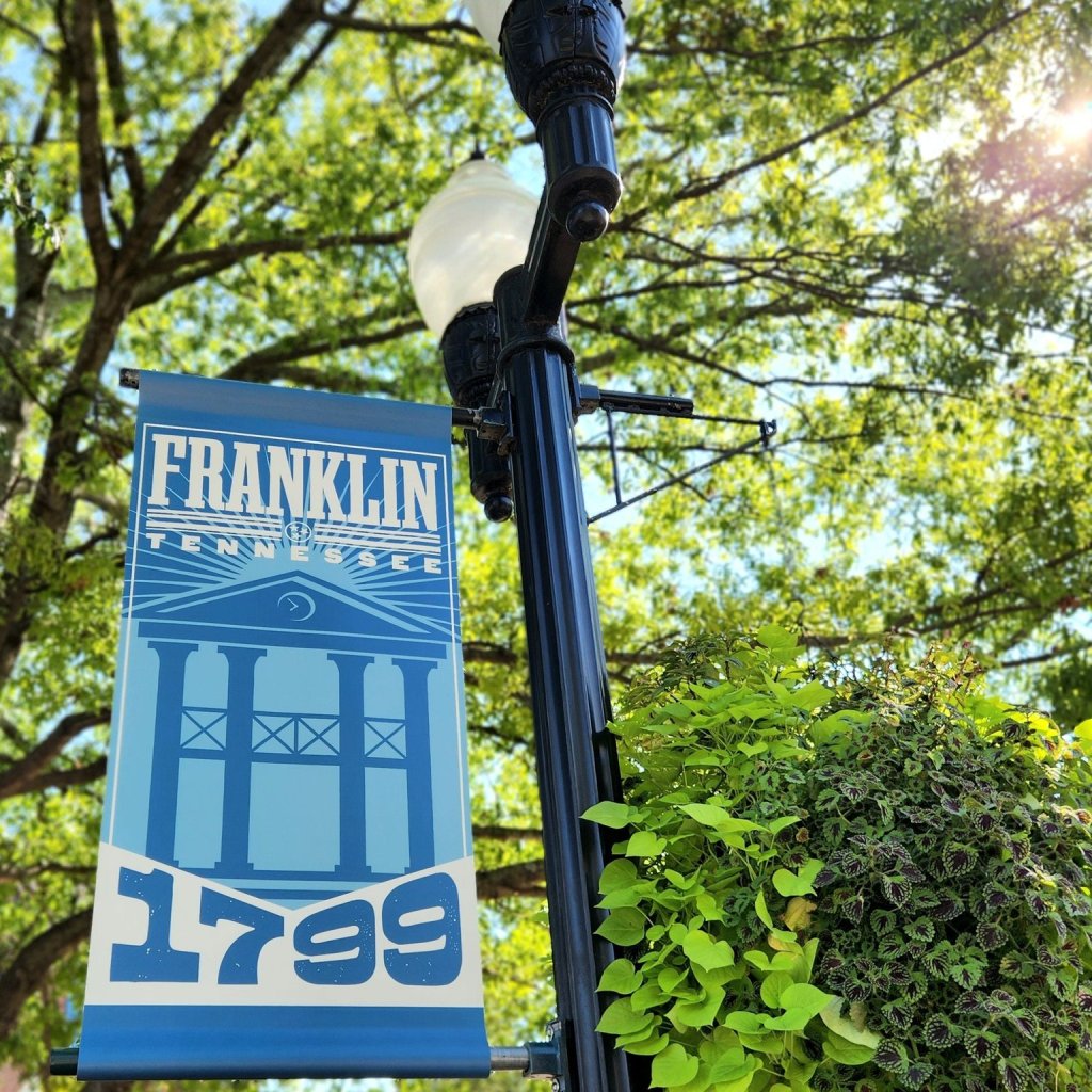 A blue banner with white text hangs from a black lamppost, surrounded by green foliage in Williamson County, Tennessee.
