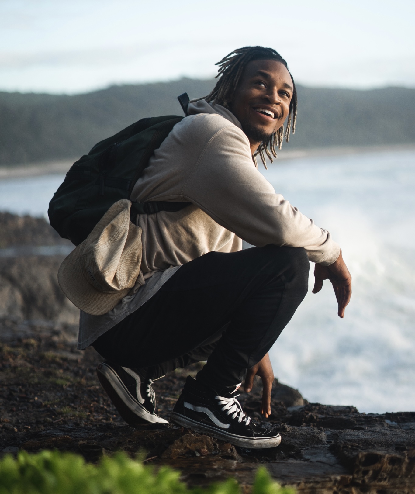 A young man with dreadlocks smiles while sitting on rocks by the ocean, wearing a backpack in Williamson County, Tennessee.