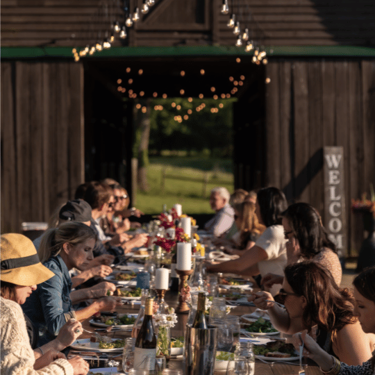 A group of people enjoys a long table dinner under a rustic wooden structure with string lights in Williamson County, Tennessee.