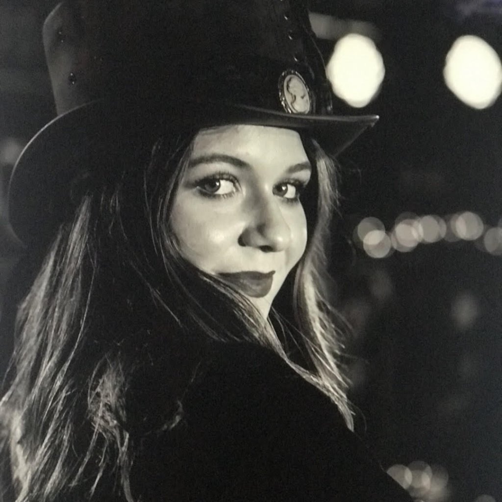 A woman with long hair smiles, wearing a black top hat with a decorative pin in Williamson County, Tennessee.