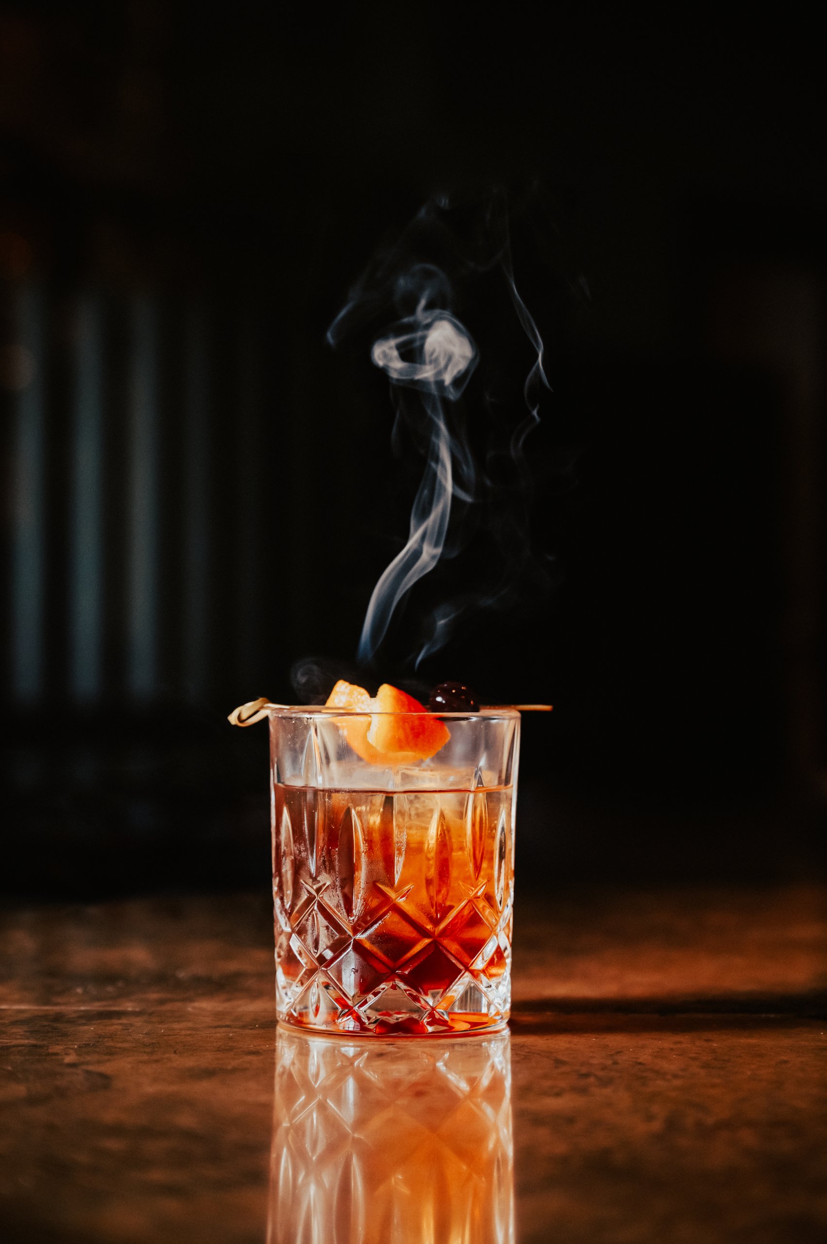 A crystal glass filled with a dark amber liquid, garnished with an orange peel and cherry, emits a plume of smoke in Williamson County, Tennessee.