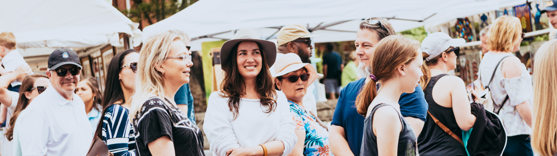 A diverse crowd of people, many wearing hats and sunglasses, gather under white tents at an outdoor market in Williamson County, Tennessee.