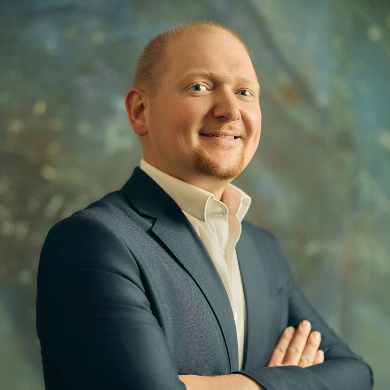 Meet The Team A smiling man in a dark suit stands confidently with arms crossed against a blue background in Williamson County, Tennessee.