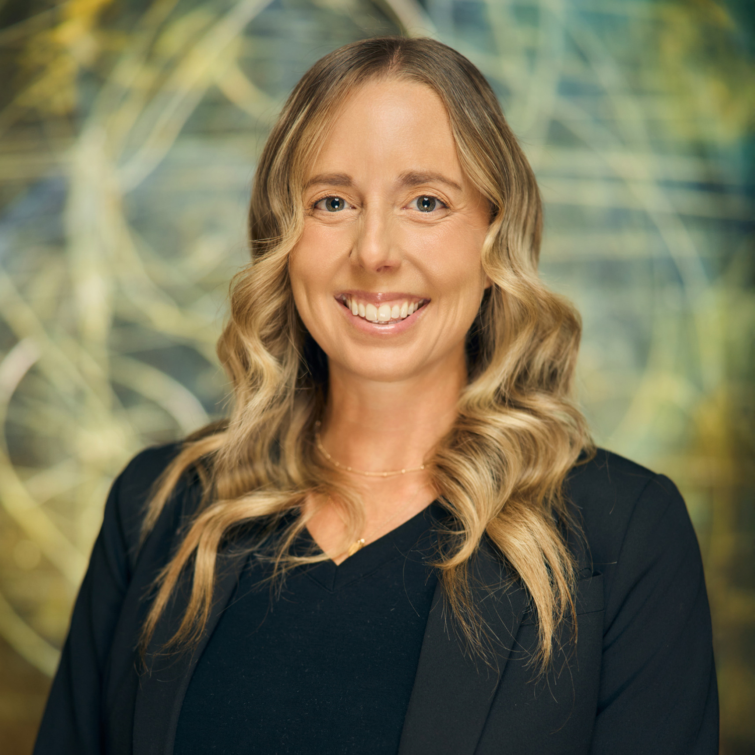 Meet The Team A smiling woman with long blonde hair wearing a black blazer stands against a colorful abstract background in Williamson County, Tennessee.