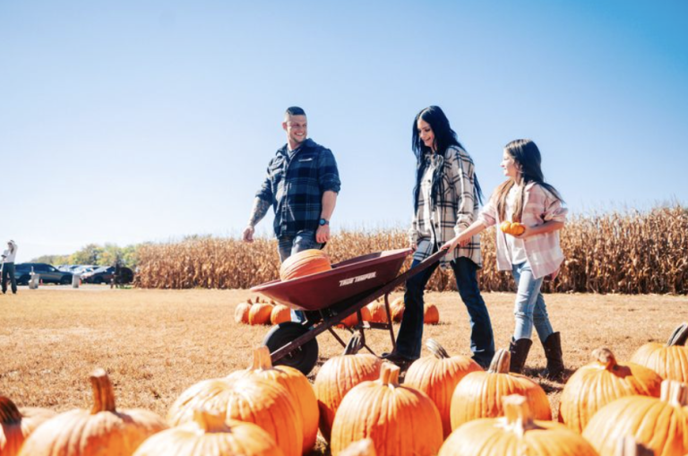 A family of three enjoys a sunny day at a pumpkin patch, pushing a wheelbarrow full of pumpkins in Williamson County, Tennessee.
