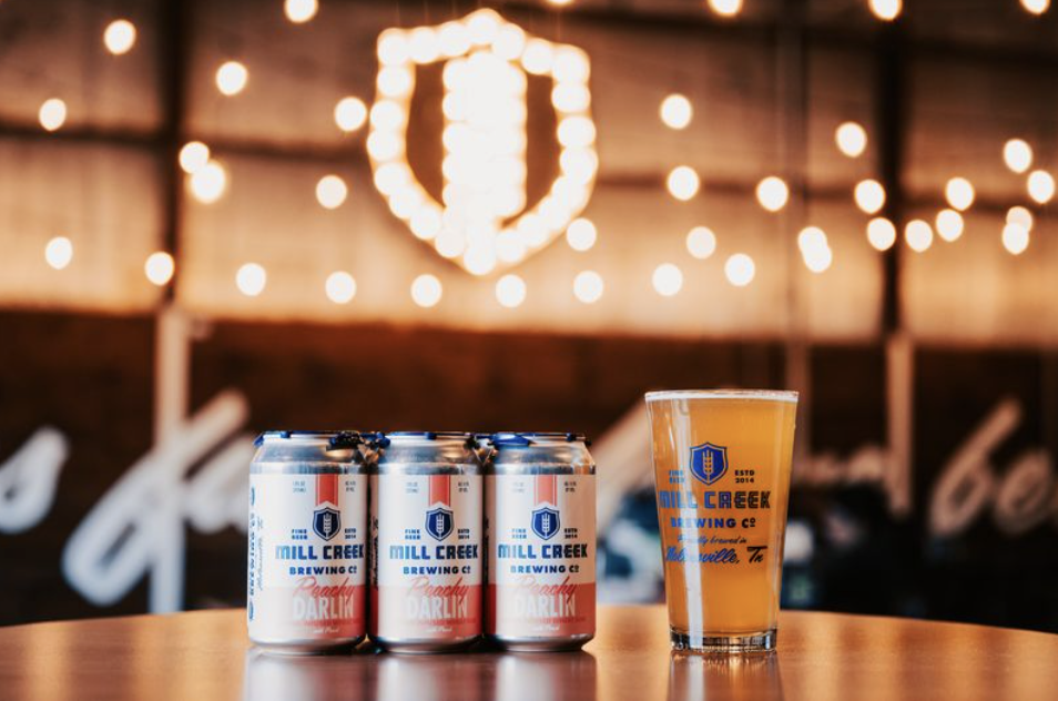 Perfect Pairings for Making the Most of Fall in Franklin A table with six cans of Mill Creek Brewing Co. beer and a glass of beer in front of a string light backdrop in Williamson County, Tennessee.