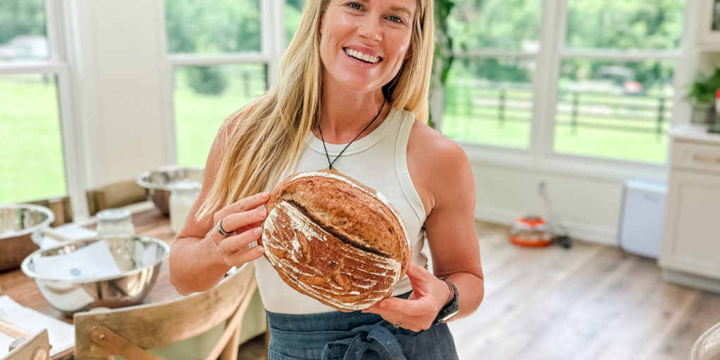 Sourdough Workshop A smiling woman holds up a round loaf of artisanal bread in a bright kitchen in Williamson County, Tennessee.