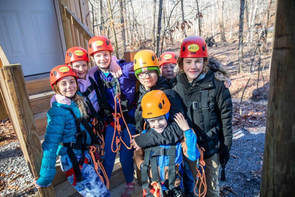 A group of eight children, all wearing colorful helmets and climbing gear, pose together on wooden stairs in a forest setting in Williamson County, Tennessee.