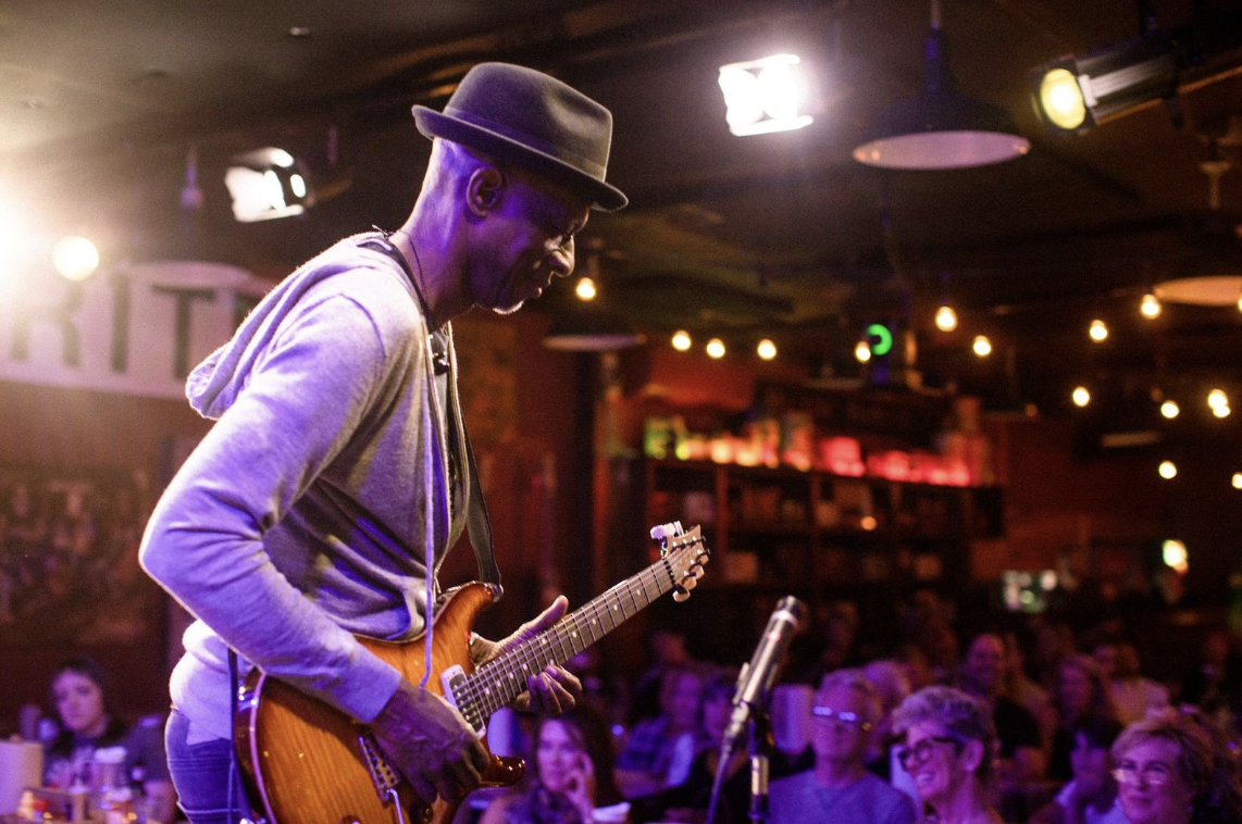 Personas Testing A musician wearing a hat plays an electric guitar on stage in a dimly lit venue in Williamson County, Tennessee.