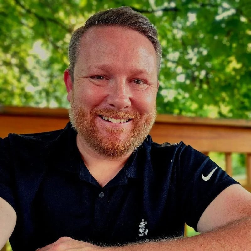 Stephen Heckler | Book Signing A smiling man with a beard wearing a black Nike shirt sits outdoors on a wooden deck in Williamson County, Tennessee.