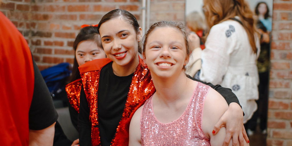 MOVE Inclusive Dance Recital: GAME ON! Three young women smile brightly, wearing sparkly outfits, at a lively indoor event in Williamson County, Tennessee.