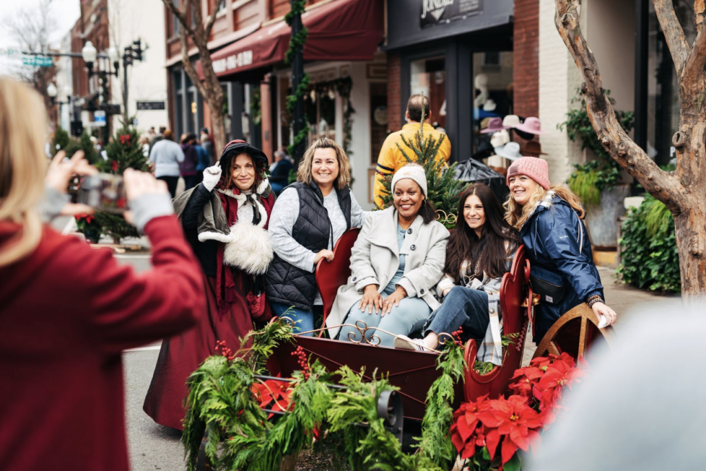 A group of six women dressed in festive winter attire pose in a decorated sleigh on a snowy street in Williamson County, Tennessee.