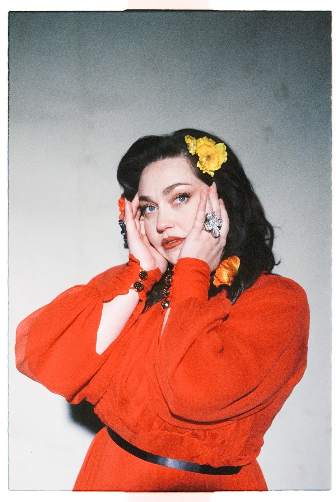 A woman in a vibrant red dress with yellow flowers in her hair touches her face with both hands in Williamson County, Tennessee.