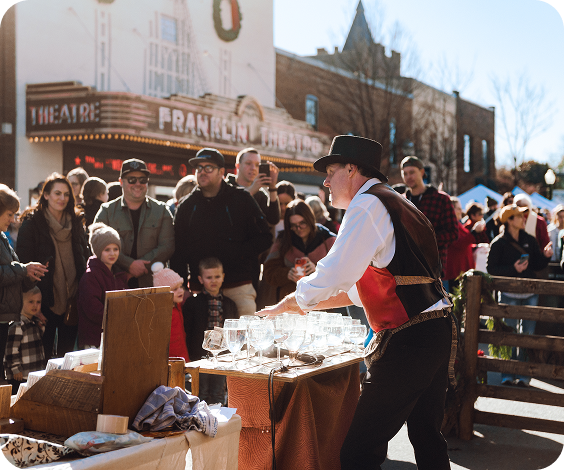 A street performer in period costume entertains a crowd with glassware tricks outside a vintage theater in Williamson County, Tennessee.