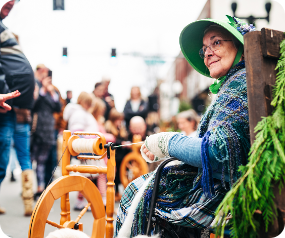 Elderly woman in traditional attire spins yarn on a wooden wheel amidst a crowd in Williamson County, Tennessee.