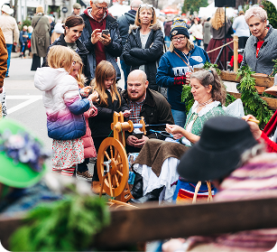 A group of people, including children, gather around a man spinning yarn on a wooden wheel at a public event in Williamson County, Tennessee.