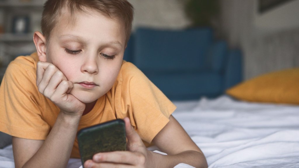 A young boy with short brown hair lies on a bed, deeply engrossed in his smartphone, his elbow propping up his head as he stares intently at the screen in Williamson County, Tennessee.