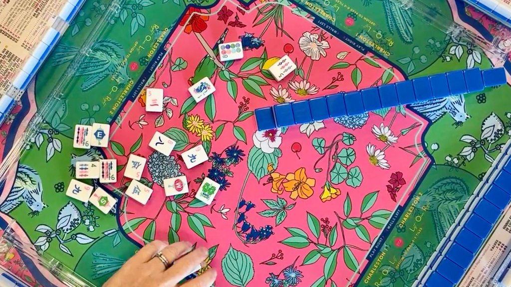 A hand reaches out to grab a blue tile on a vibrant floral-patterned Mahjong board with scattered tiles in Williamson County, Tennessee.