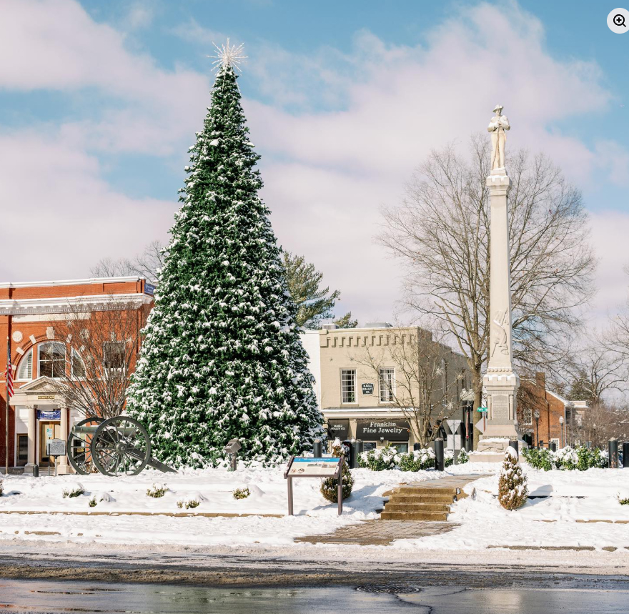 A picturesque town square features a towering Christmas tree adorned with lights and topped with a star, standing tall amidst a blanket of fresh snow, with a historic monument and quaint buildings in the background in Williamson County, Tennessee.