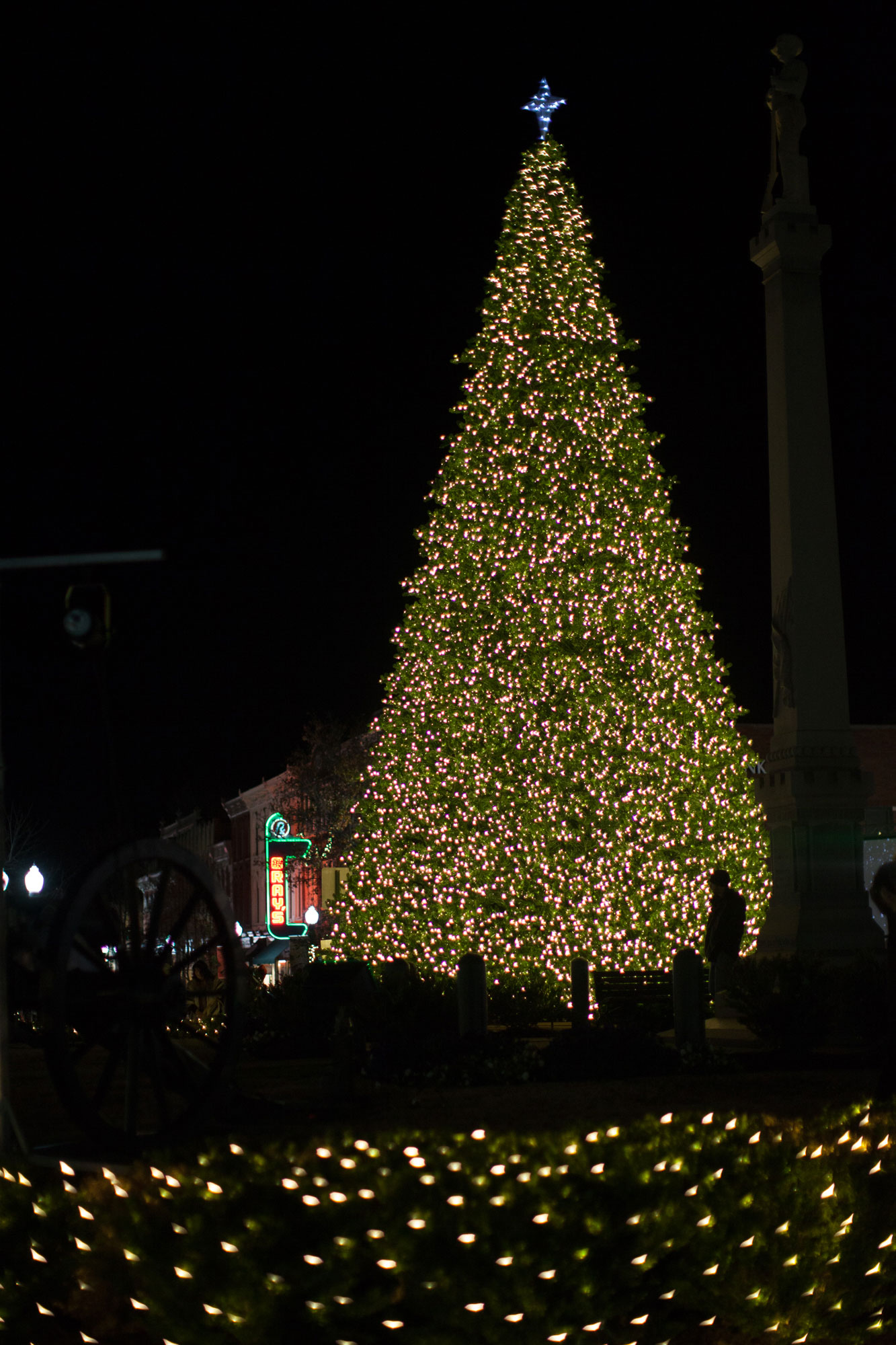 A majestic Christmas tree, adorned with countless twinkling lights, stands tall against the night sky, with a statue and illuminated signs in the background in Williamson County, Tennessee.