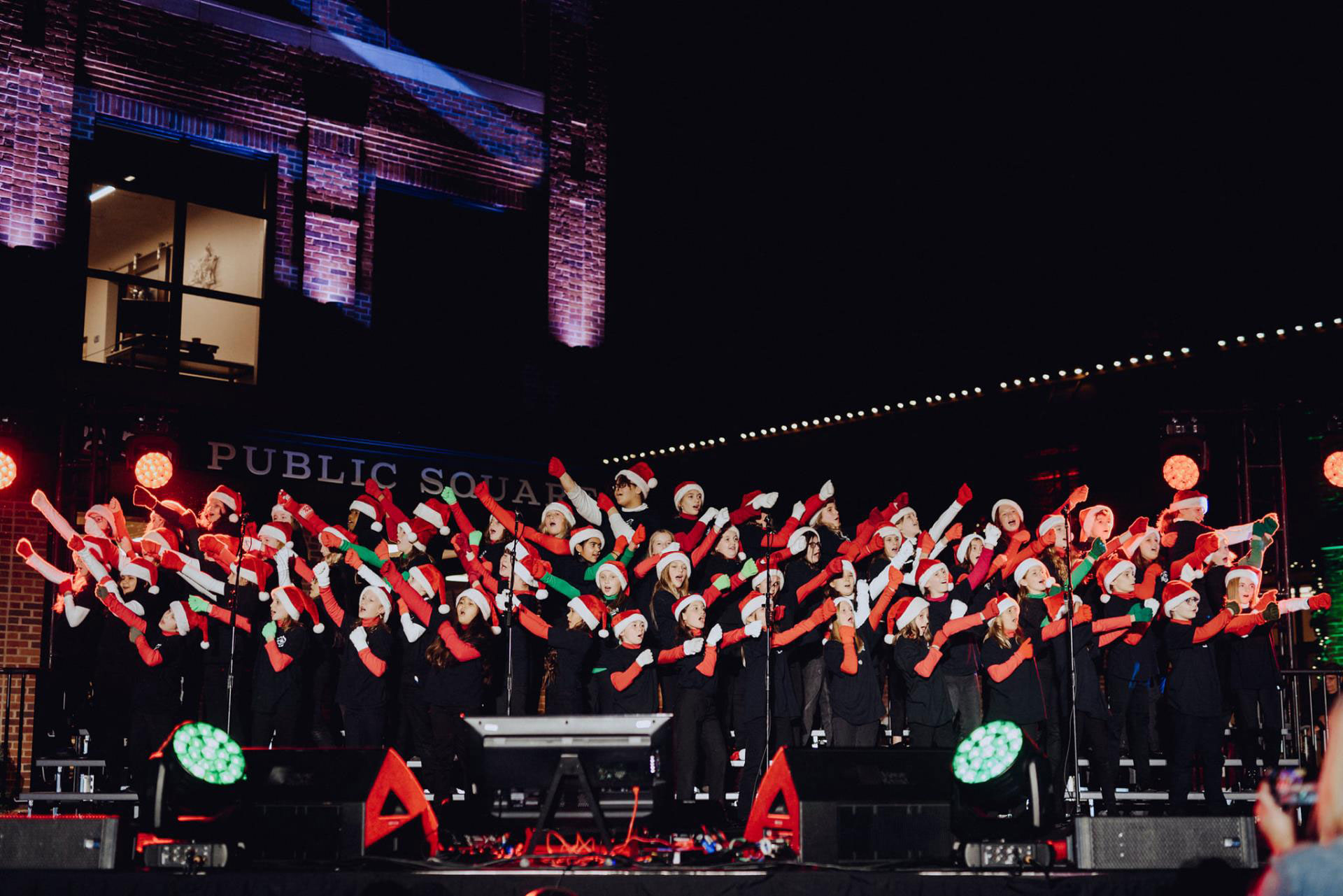 A large choir dressed in black with red and white Santa hats performs on a stage illuminated by festive lights, singing with enthusiasm and joy in Williamson County, Tennessee.