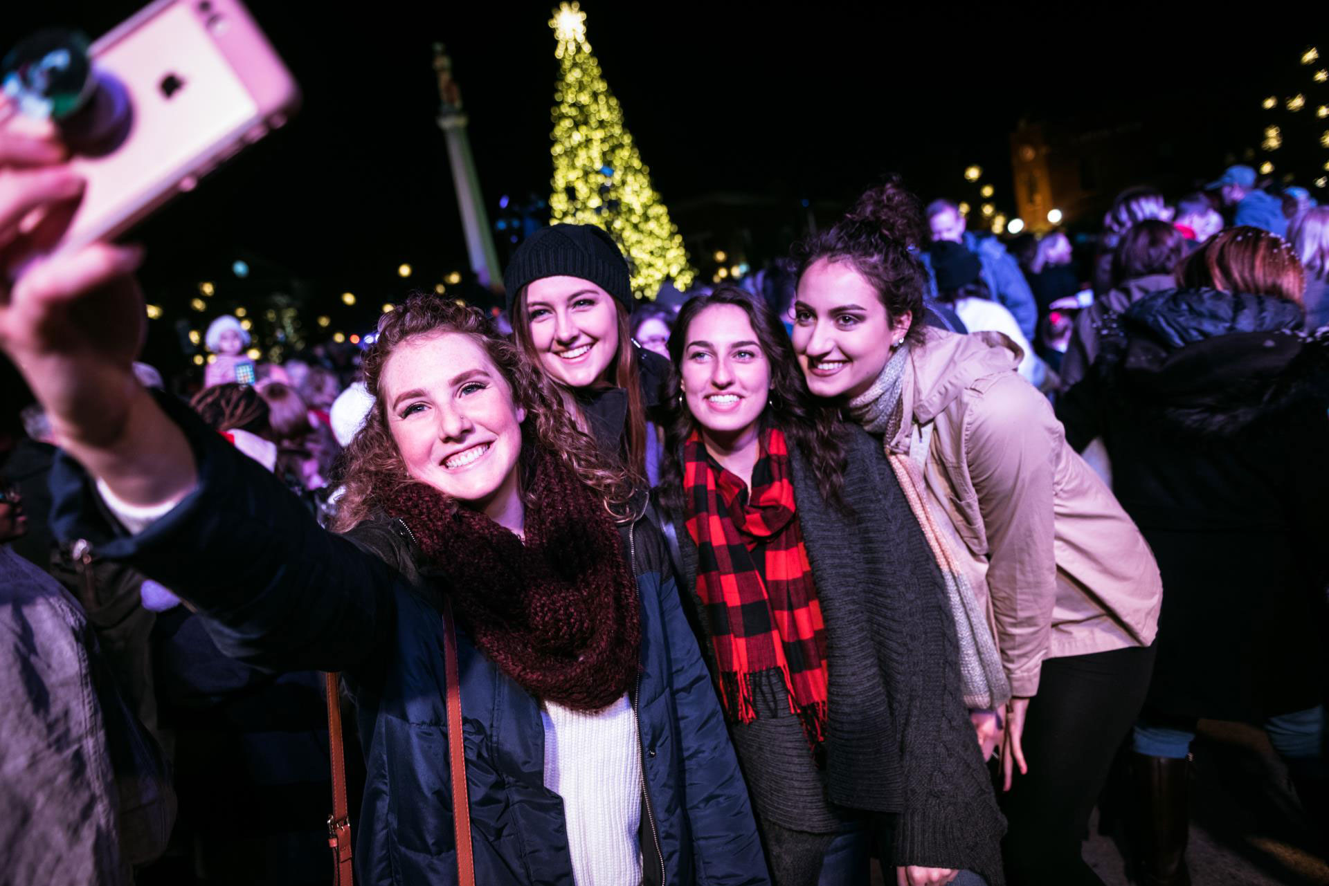 Four women take a selfie in front of a brightly lit Christmas tree, surrounded by a festive crowd in Williamson County, Tennessee.