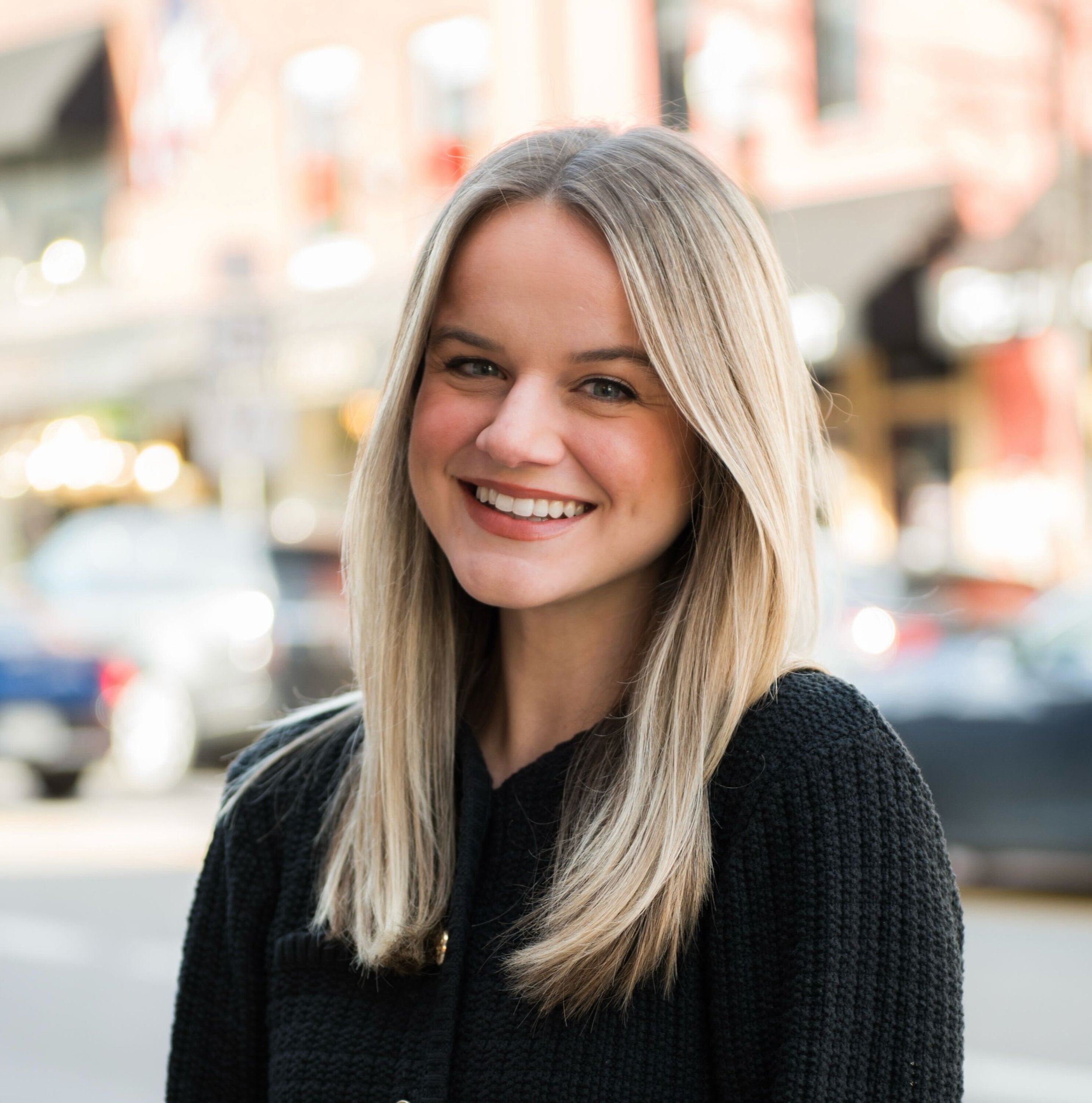 A smiling woman with long blonde hair wearing a black sweater stands on a city street in Williamson County, Tennessee.