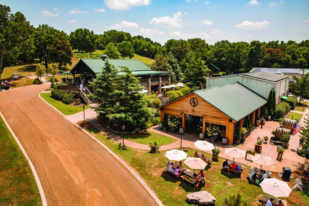 A picturesque outdoor venue features a rustic wooden building with a green roof, surrounded by lush greenery, picnic tables with umbrellas, and a winding dirt road in Williamson County, Tennessee.