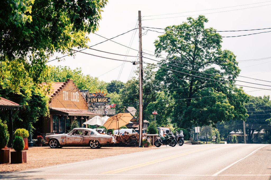 A vintage car is parked outside a rustic music venue with a sign advertising live music tonight, surrounded by lush greenery and a quiet road in Williamson County, Tennessee.