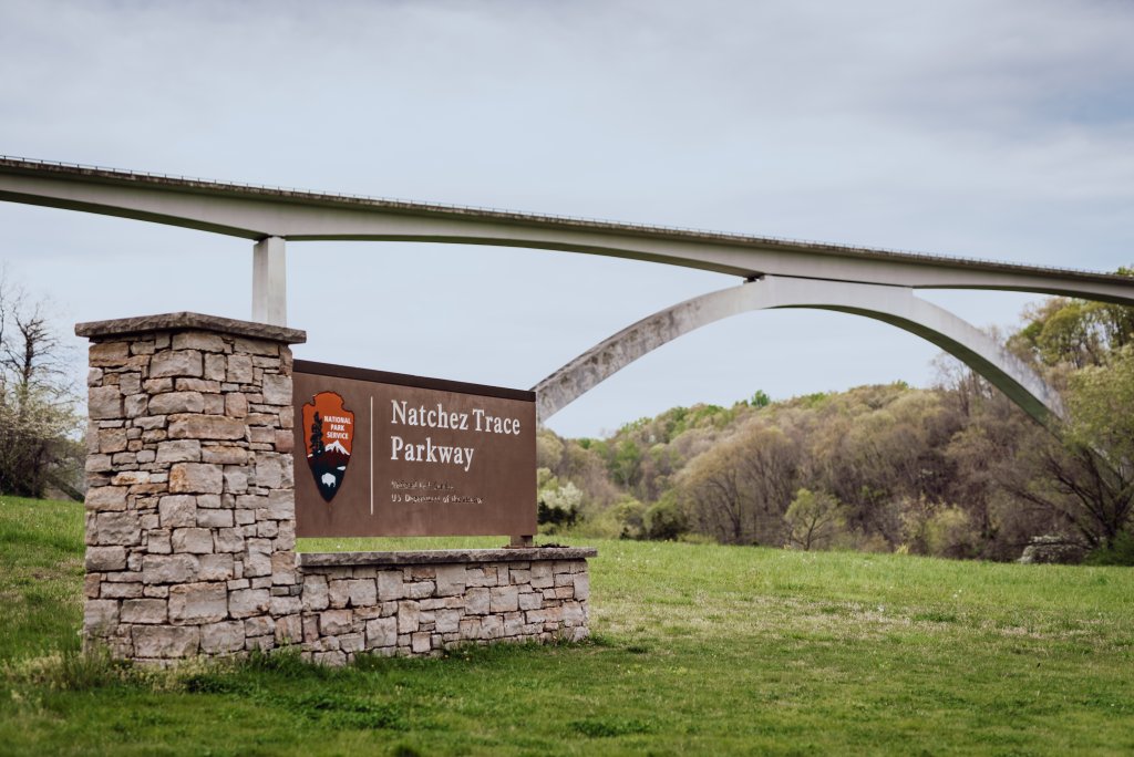 A stone sign with the National Park Service logo marks the entrance to Natchez Trace Parkway, with a graceful arch bridge spanning the background in Williamson County, Tennessee.