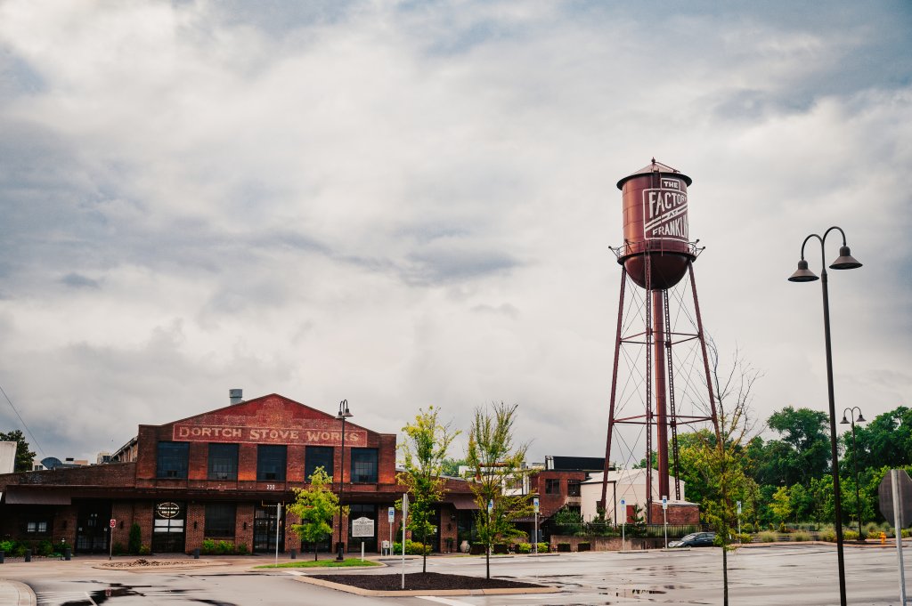 A historic red brick building with the sign 'Dortch Stove Works' and a tall water tower with 'The Factory at Franklin' painted on it stand under a cloudy sky in Williamson County, Tennessee.