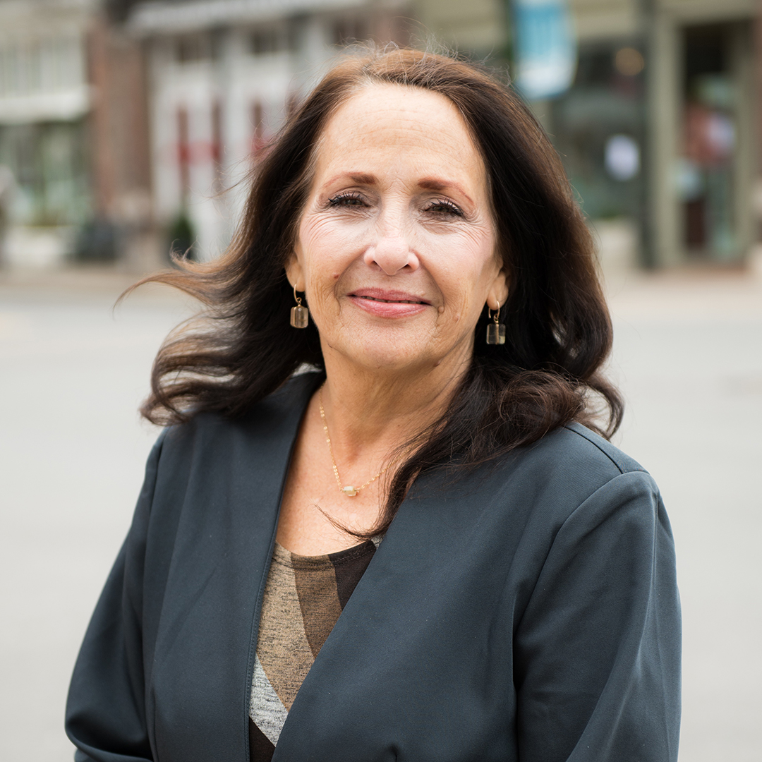 A woman with long, dark hair is smiling warmly at the camera, wearing a dark blazer over a striped top, with a necklace and earrings in Williamson County, Tennessee.