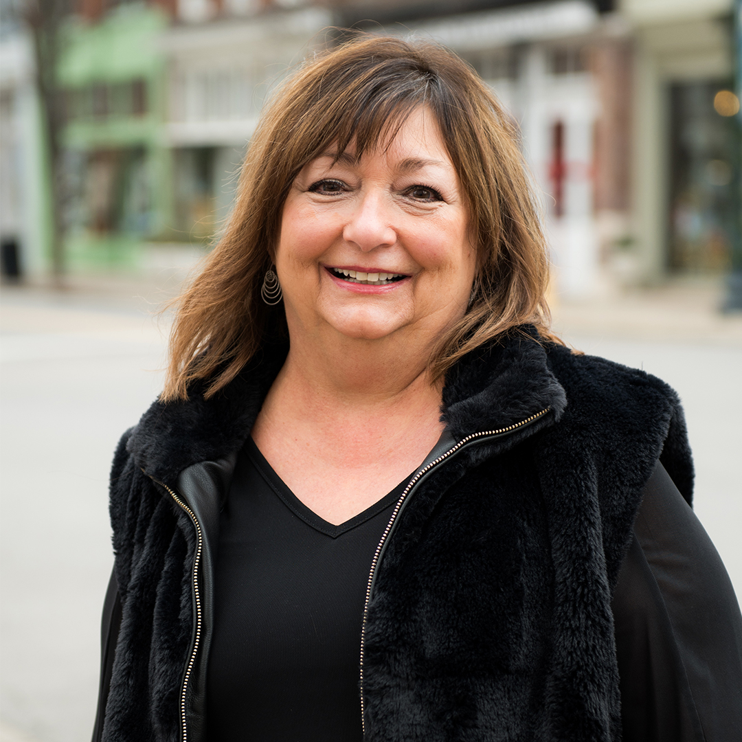 A smiling woman with shoulder-length brown hair wearing a black furry vest over a black top stands outdoors on a city street in Williamson County, Tennessee.
