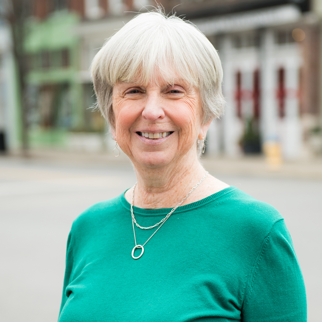 A smiling woman with short white hair wearing a green sweater and a silver necklace stands outdoors in Williamson County, Tennessee.