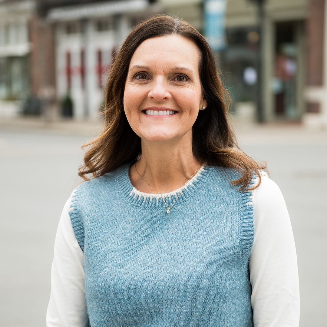 A woman with long, wavy brown hair is smiling warmly at the camera, wearing a blue sweater vest over a white long-sleeved shirt in Williamson County, Tennessee.
