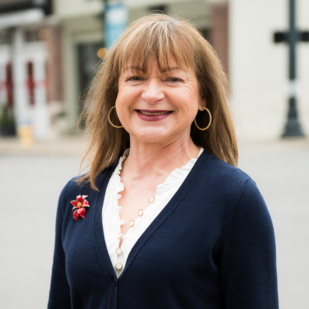 A woman with shoulder-length brown hair is smiling warmly, wearing a navy blue cardigan with a red flower brooch, a white blouse, and gold hoop earrings in Williamson County, Tennessee.
