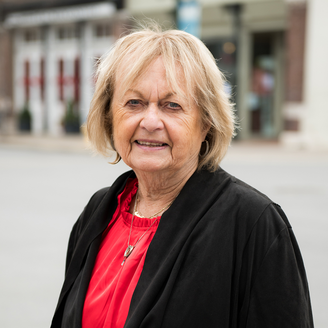 A smiling woman with short blonde hair wearing a black cardigan over a red blouse stands outdoors in front of a building with red doors in Williamson County, Tennessee.