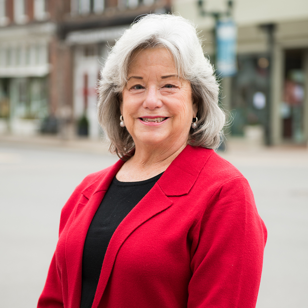 A woman with short, gray hair is smiling warmly at the camera, wearing a bright red cardigan over a black top in Williamson County, Tennessee.