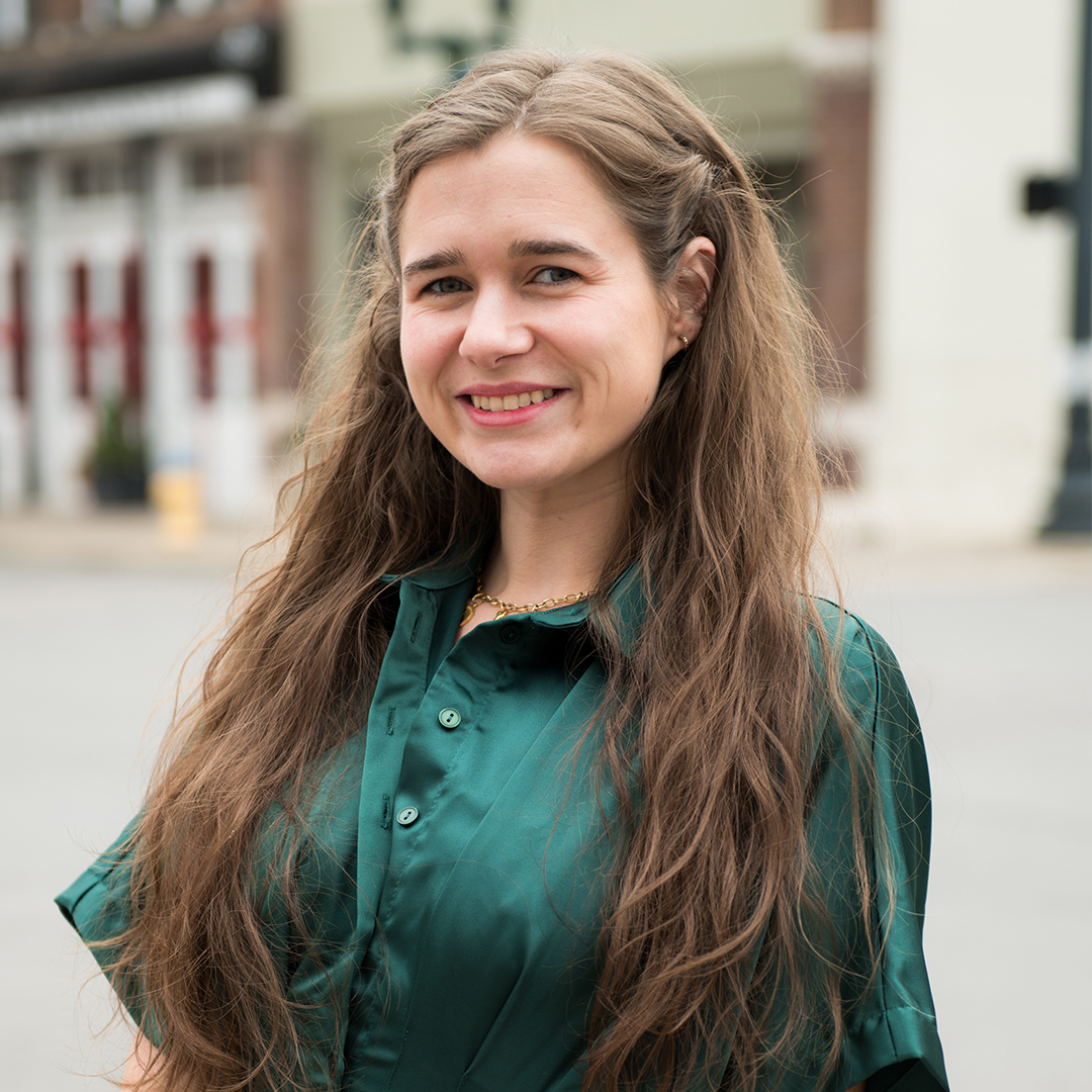 A woman with long, wavy brown hair is smiling warmly at the camera, wearing a green button-up shirt and a gold necklace in Williamson County, Tennessee.