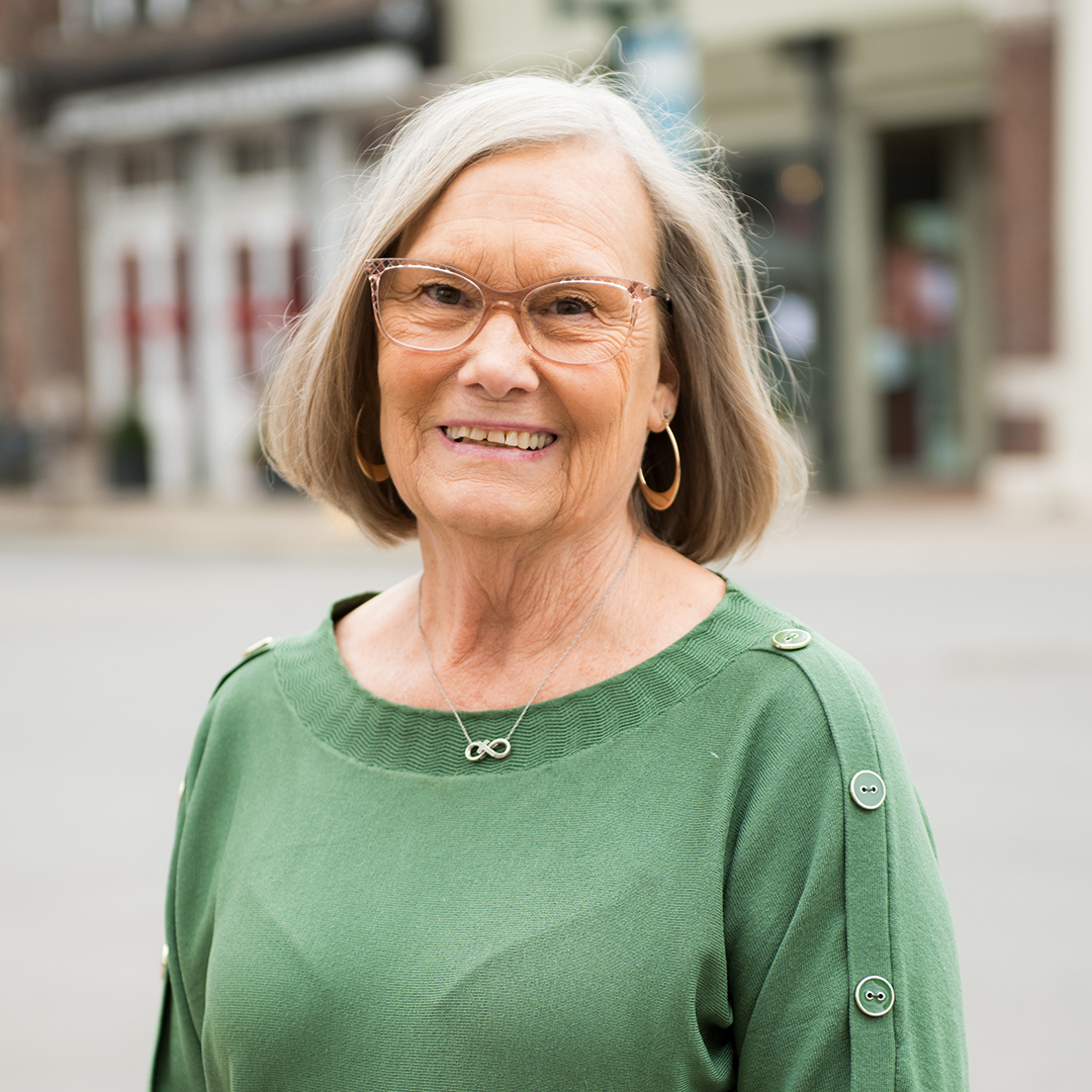 A smiling elderly woman with short gray hair and glasses wearing a green sweater with buttons and a necklace with an infinity symbol pendant in Williamson County, Tennessee.