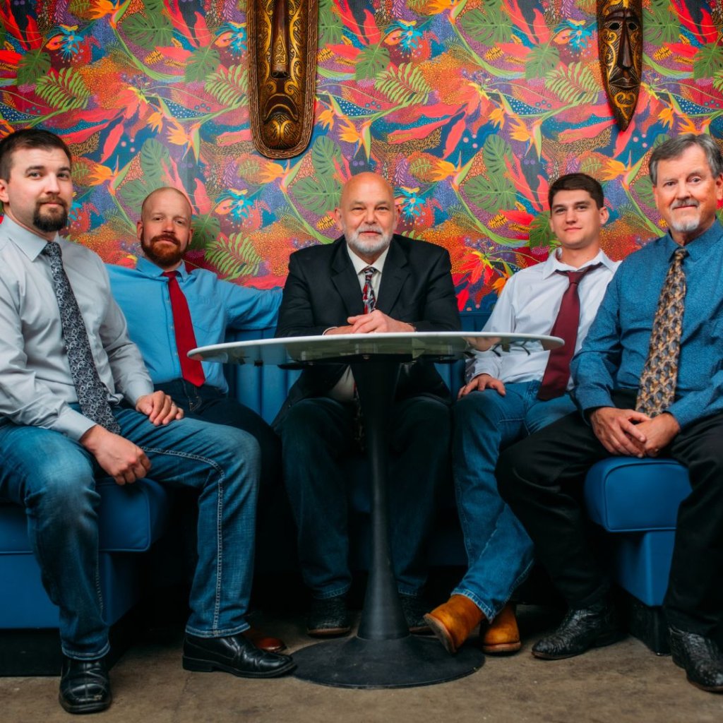 Five men dressed in business casual attire sit on a blue couch against a vibrant, colorful tropical wallpaper backdrop in Williamson County, Tennessee.