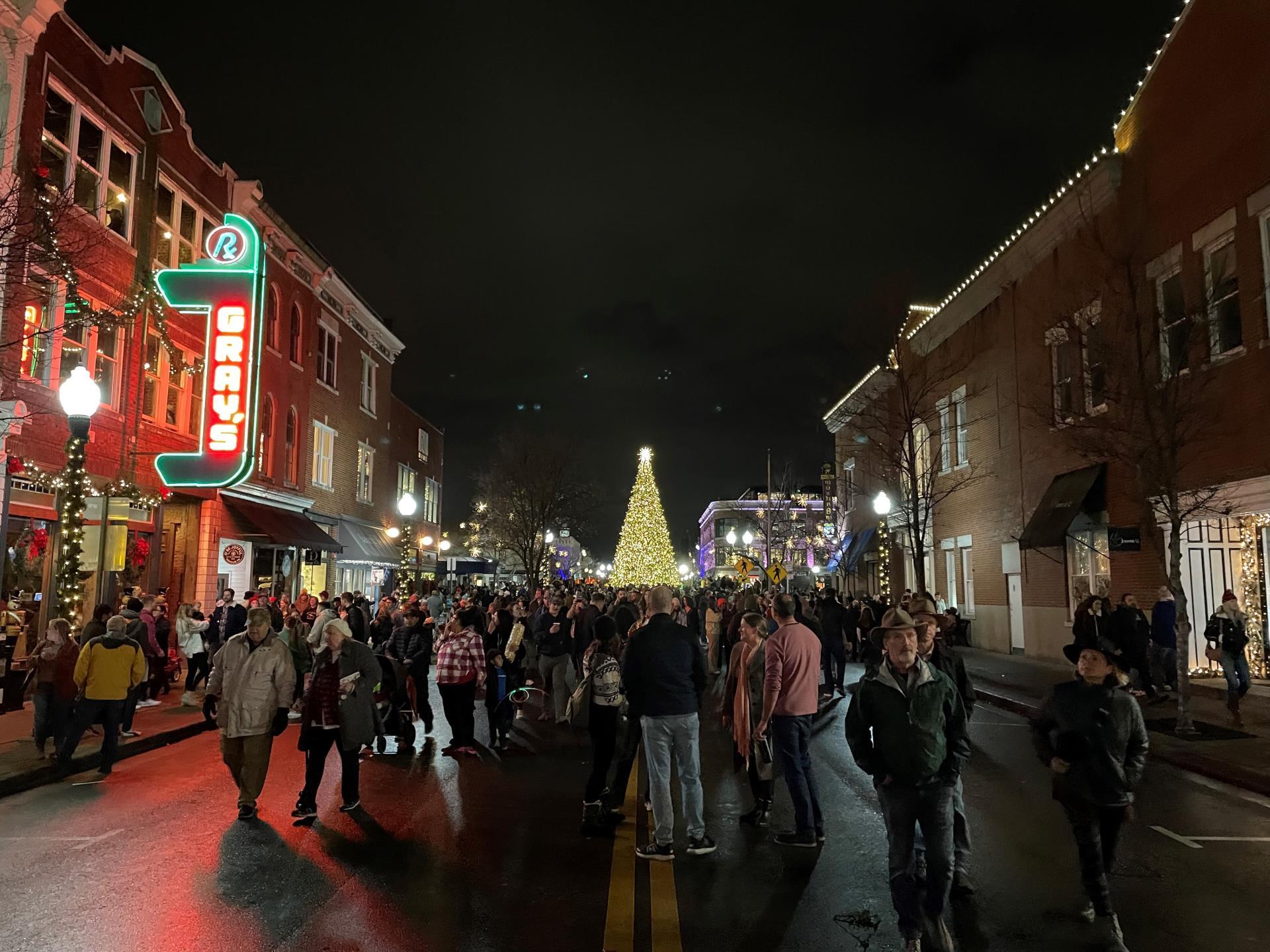 A bustling night scene on a wet street during a Christmas festival, with a large crowd gathered to admire a towering illuminated tree and festive storefronts in Williamson County, Tennessee.