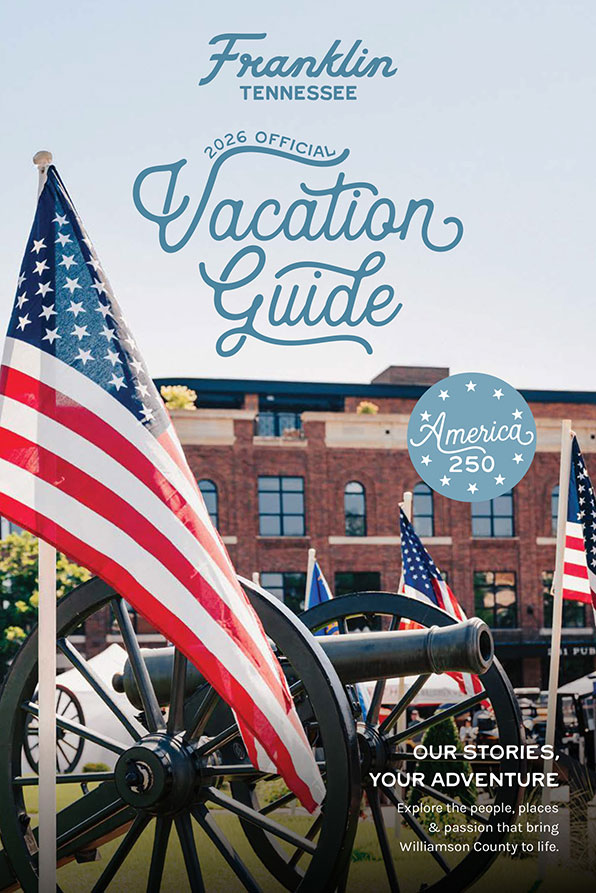 2023-Official-Vacation-Guide-DIGITAL-1 A vibrant American flag waves proudly in front of a historic cannon, set against the backdrop of a charming brick building, inviting visitors to explore Williamson County's rich history and vibrant culture in Williamson County, Tennessee.