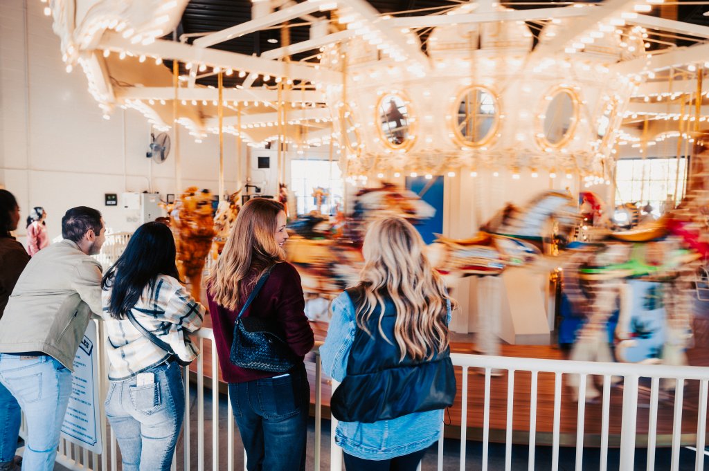 A group of people, including two women with long blonde hair, are standing at the railing of a beautifully lit carousel, watching the colorful horses and lights spin around in Williamson County, Tennessee.